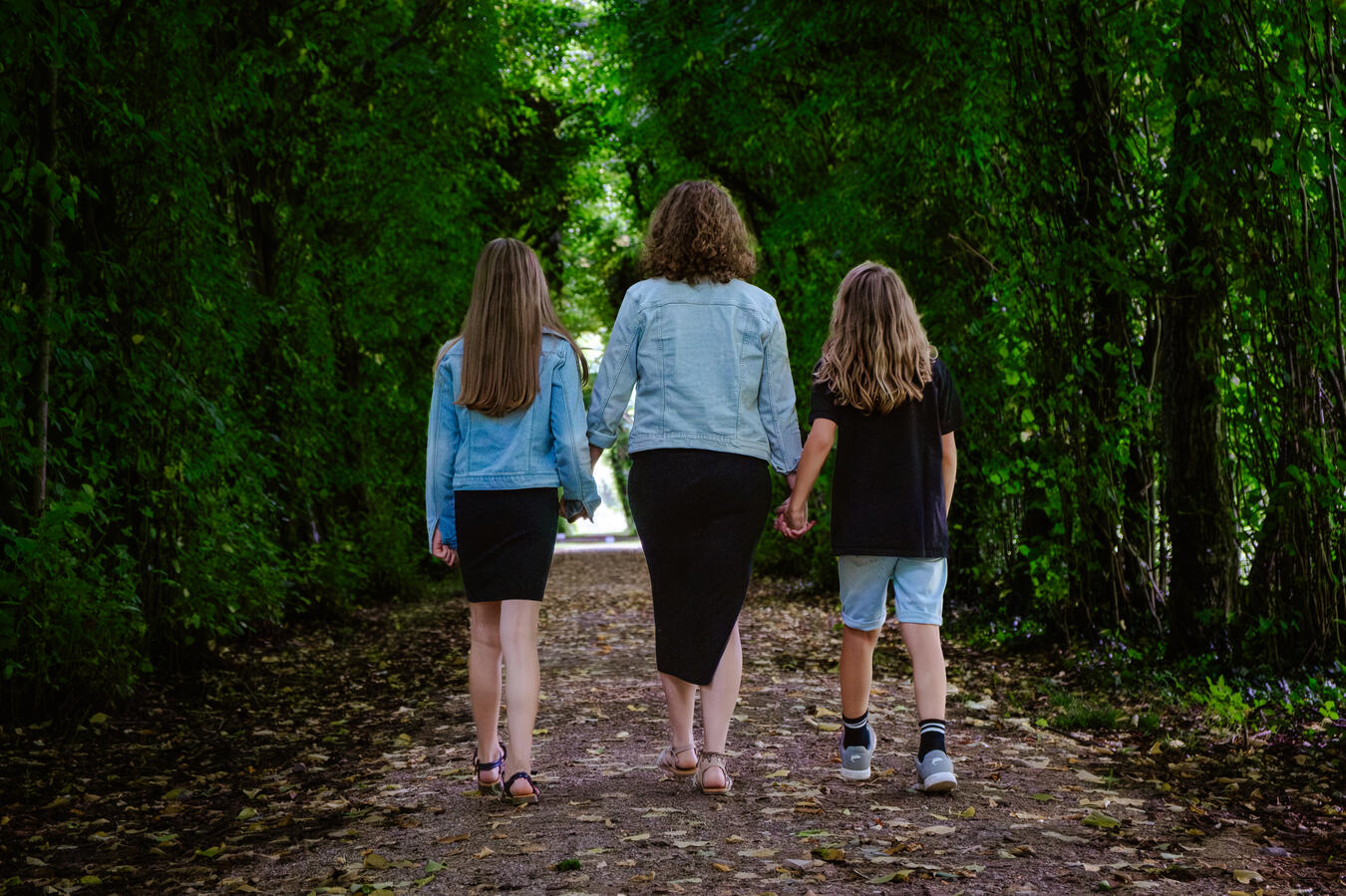 Farbaufnahme von Mutter mit zwei Kindern, die gemeinsam auf einem von Bäumen gesäumten Waldweg gehen.