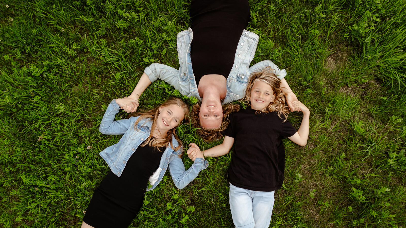 Drohnenaufnahme einer Mutter mit zwei Kindern, Hand in Hand liegend auf einer grünen Wiese – Familienfoto aus der Luft