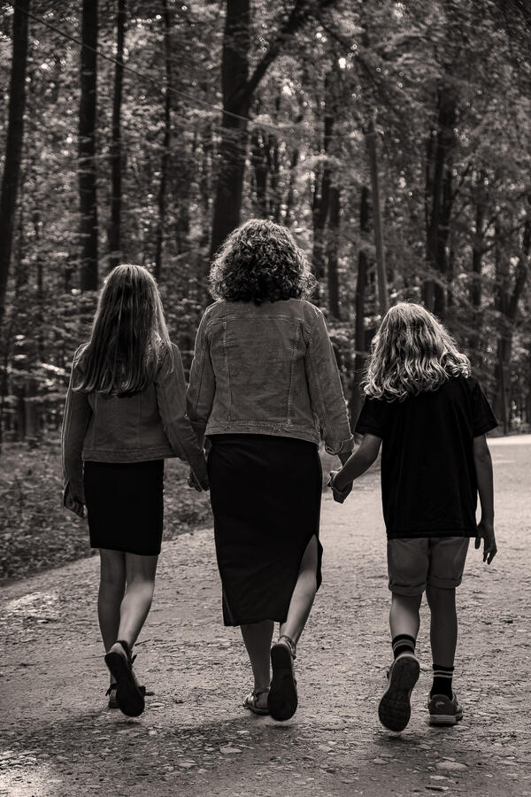 Mutter und zwei Kinder gehen Hand in Hand auf einem Waldweg – Schwarzweißaufnahme von hinten.