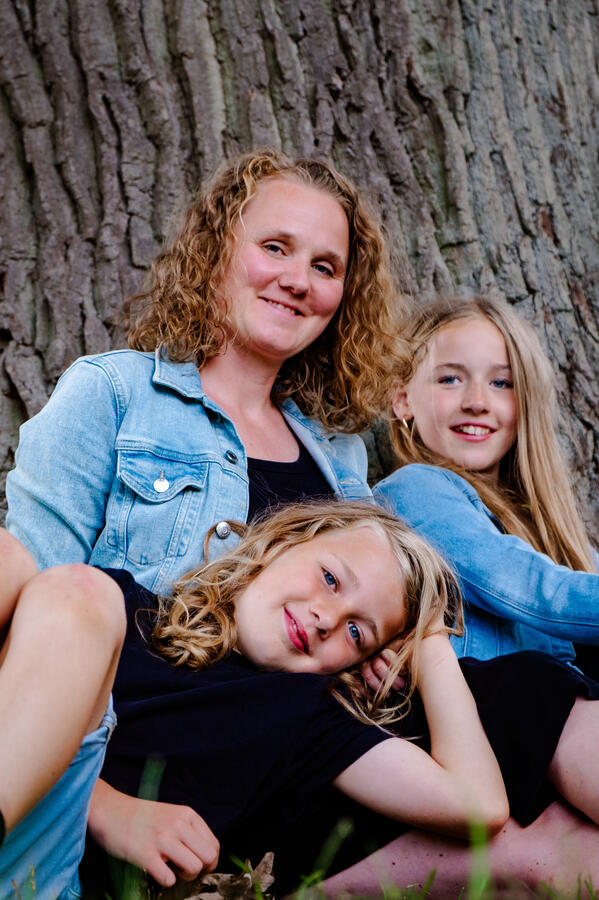 Mutter mit zwei Kindern an einem Baum lehnend, alle lächeln – natürliches Familienfoto im Freien