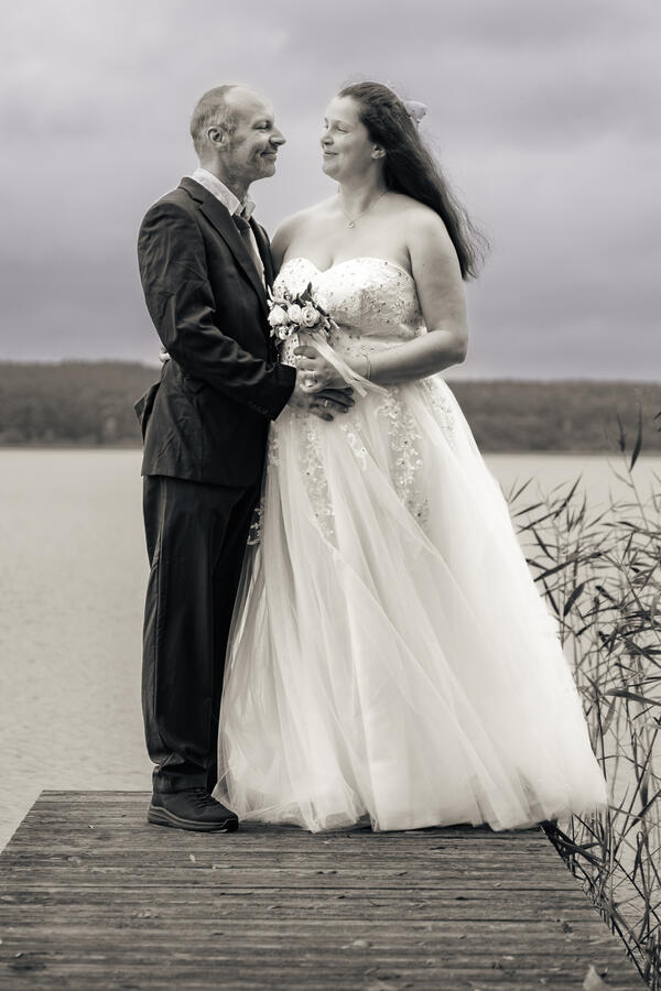Hochzeitspaar steht eng beisammen am Wasser; die Braut hält ihren Strauß und das Kleid fällt weich im Wind.