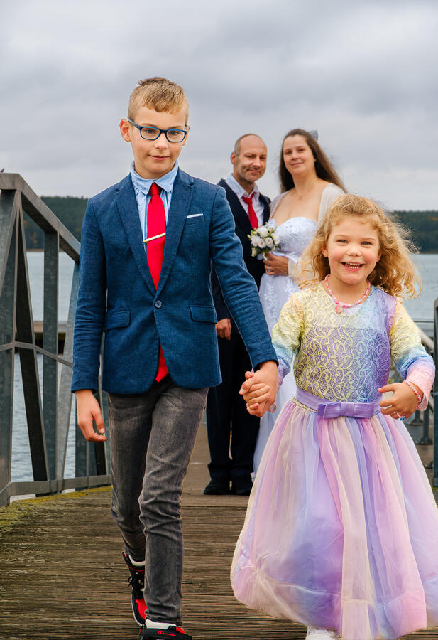 Lachendes Kind in festlichem Kleid läuft an der Hand einer erwachsenen Person über eine Brücke, begleitet von Hochzeitsgästen im Hintergrund.
