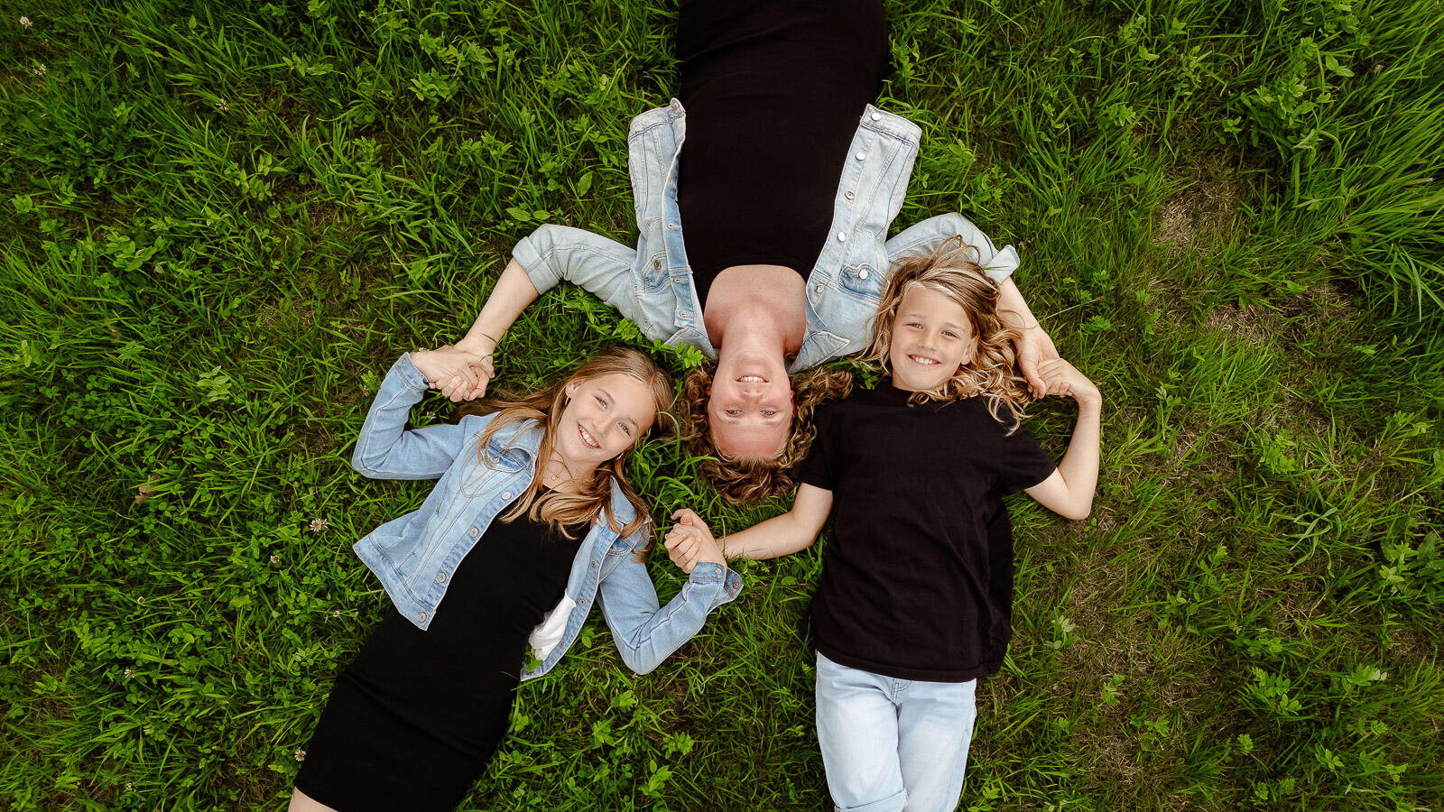 Drohnenfoto von Mutter mit zwei Kindern, liegend auf grüner Wiese und Hand in Hand – emotionale Luftaufnahme einer Familie