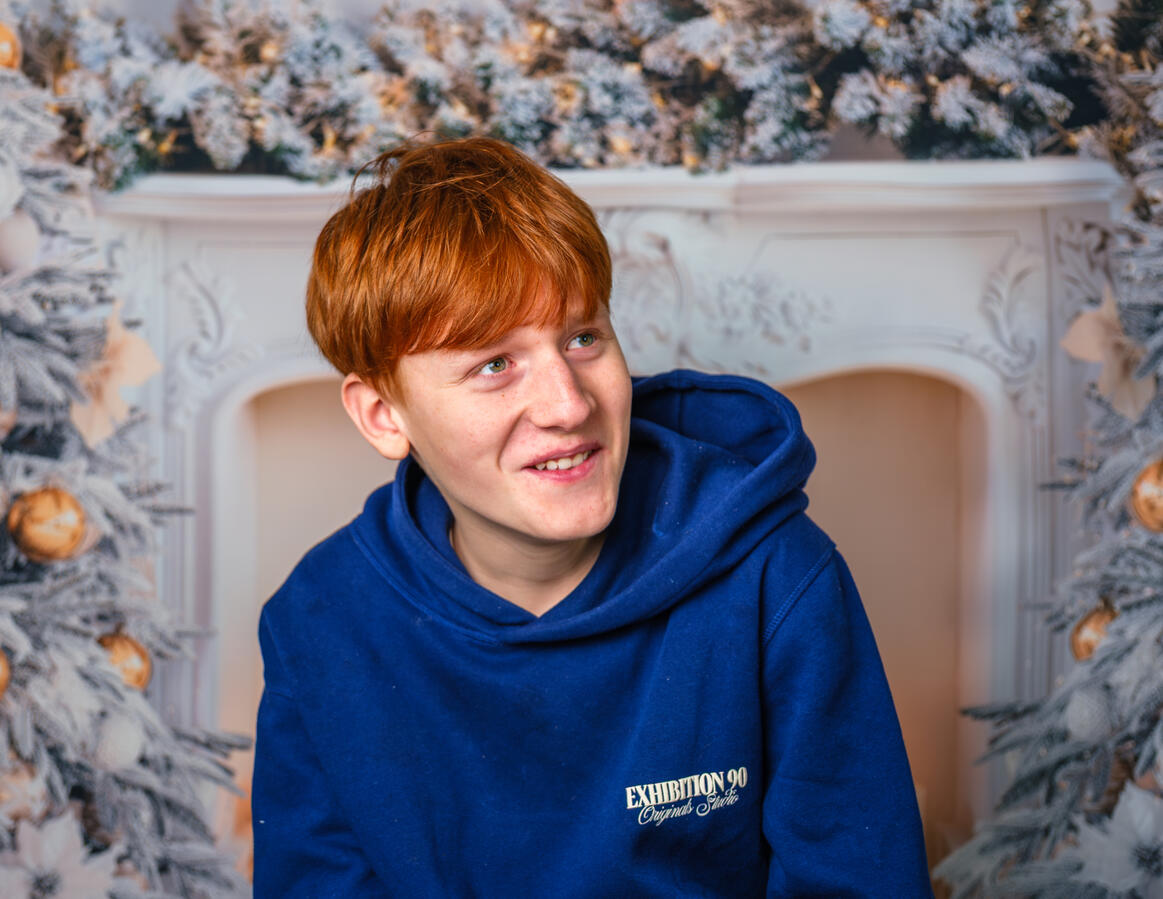 Portrait eines Jungen in blauem Pullover vor einer winterlichen Hintergrundkulisse. Ein natürlicher, ruhiger Moment aus einem Familienshooting, aufgenommen in weichem, stimmungsvollem Licht.