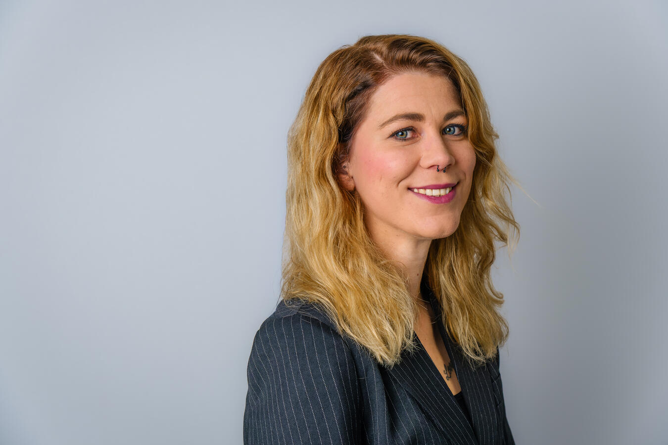 Ein natürliches Portrait einer jungen Frau mit lockigem blondem Haar vor neutralem Hintergrund. Das Bild zeigt einen freundlichen, authentischen Ausdruck und wurde mit weichem, professionellem Licht aufgenommen – ideal für Business- oder Lifestyle-Darstell