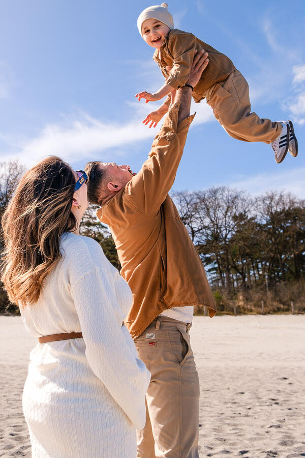 Actionreiches Familienfoto am Strand: Ein lachender Vater wirft sein Kind spielerisch in die Luft, während die Mutter glücklich daneben steht