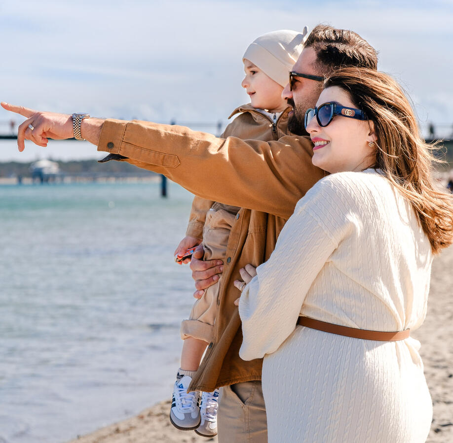 Emotionale Familienfotografie am Timmendorfer Strand: Vater hält Kind auf dem Arm, während die schwangere Mutter lächelnd mit ihnen in die Ferne blickt.