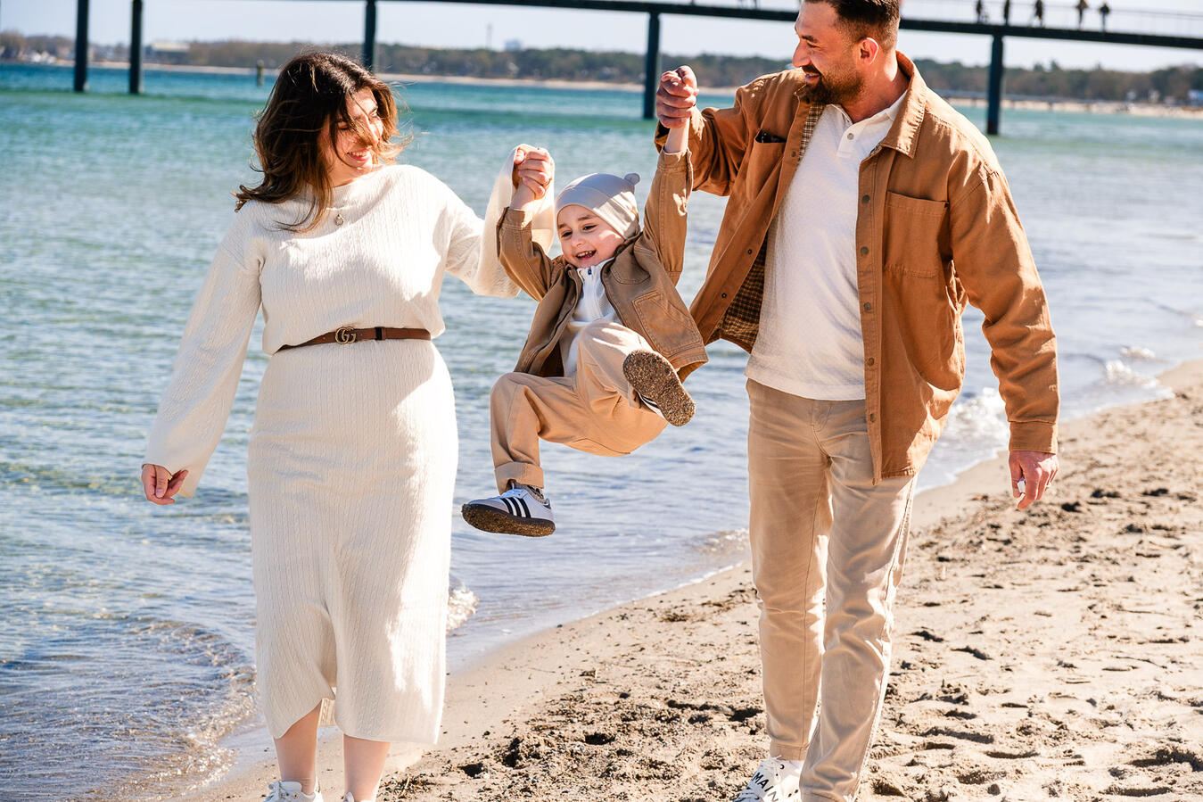 Fröhliches Familienshooting am Strand: Eltern lassen ihr lachendes Kind in die Luft fliegen, im Hintergrund die Seebrücke und das Meer.