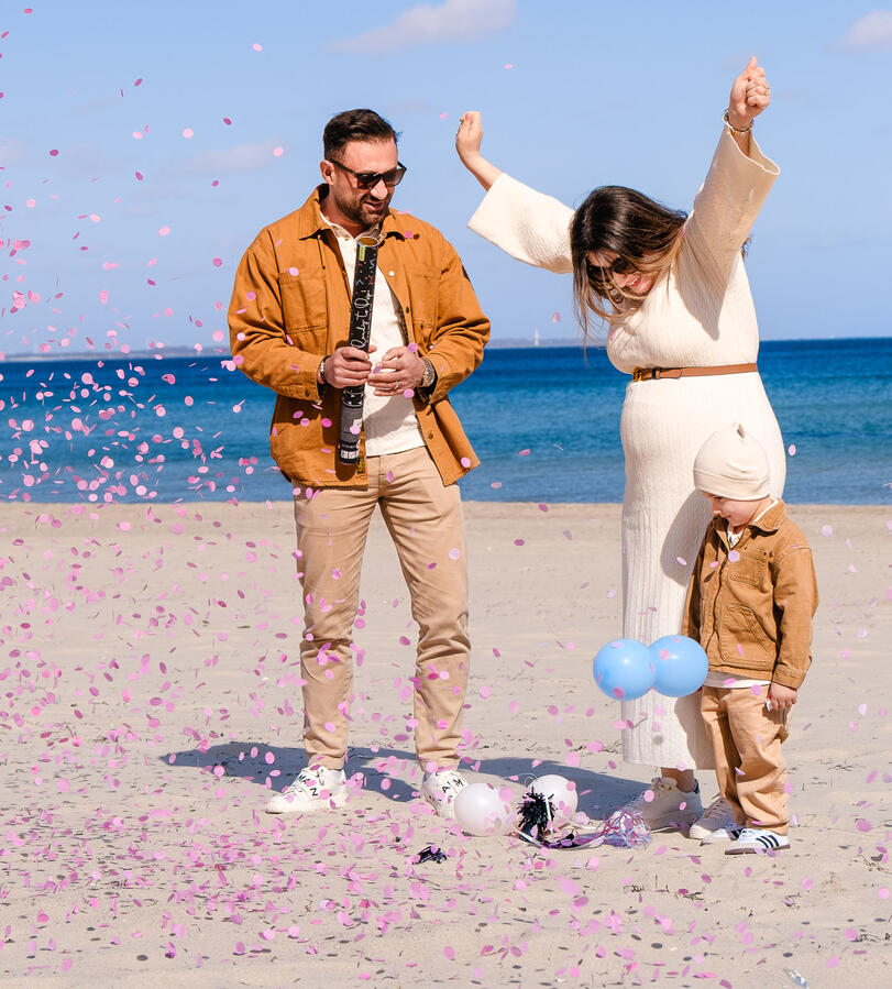 Gender Reveal Shooting am Strand: Eine Familie feiert mit einer Konfetti-Kanone und rosa Konfetti im Wind, während blaue Luftballons im Sand liegen.
