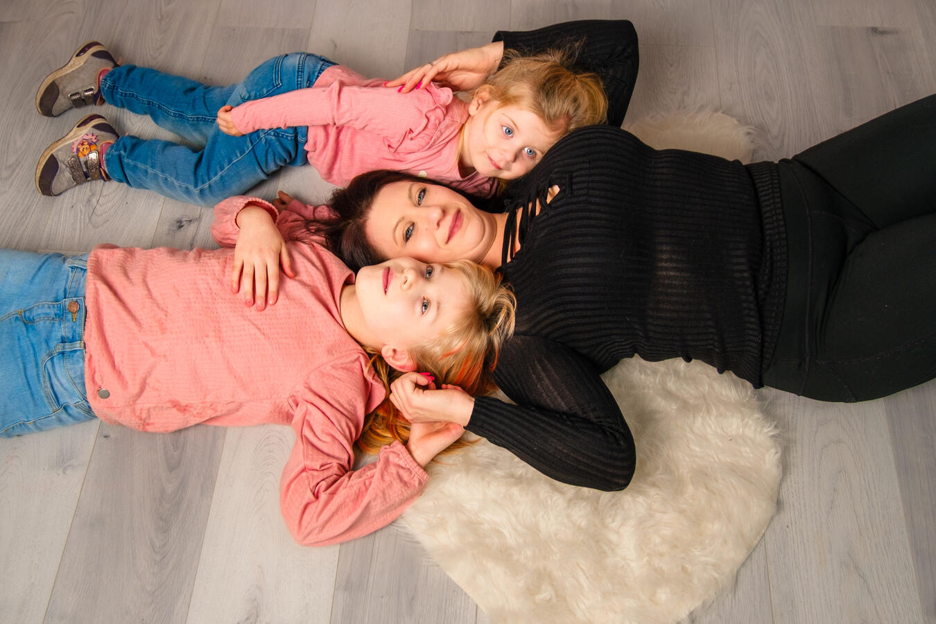 Drei Personen liegen gemeinsam entspannt auf einem hellen Boden mit einem Fellteppich und bilden eine harmonische Familienszene.