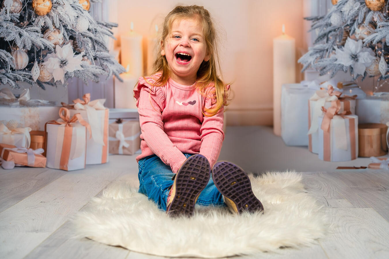 Lachendes Mädchen sitzt auf einem weißen Fellteppich in einer weihnachtlich dekorierten Szene mit Geschenken und Lichterdekoration.